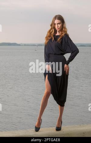 Attraktives Mädchen, das ein langes schwarzes Kleid trägt und seine schlanken Beine am Pier zeigt Stockfoto