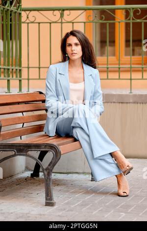 Stilvolle Frau in einem himmelblauen Hosenanzug, die sich auf einer Bank in der Nähe des Büros ausruhen kann. Stockfoto