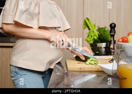 Nahaufnahme der Hände einer nicht erkennbaren Schwangeren in der Küche, die frische und saftige Gurken für den Salat schneiden Stockfoto