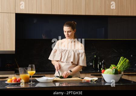 Eine Schwangerin in der Küche schneidet frische und saftige Gurken für den Salat Stockfoto
