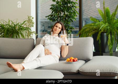 Die süße Schwangere sitzt mit einem Glas frischen Orangensaft in der Hand auf dem Sofa Stockfoto