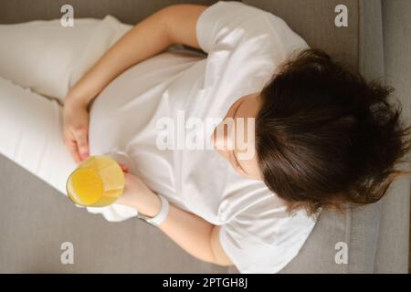 Draufsicht auf eine Schwangere, die mit einem Glas frischen Orangensaft in der Hand auf dem Sofa sitzt Stockfoto