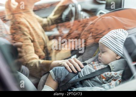 Mutter konzentriert sich darauf, mit dem Familienauto Besorgungen zu machen, während ihr Baby auf dem Kindersitz neben ihrem Standort schläft Stockfoto