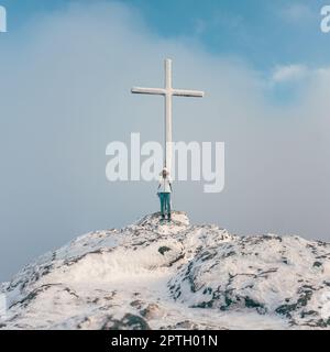 Frau, die in der Nähe des Gipfelkreuzes auf einem Berggipfel steht Stockfoto