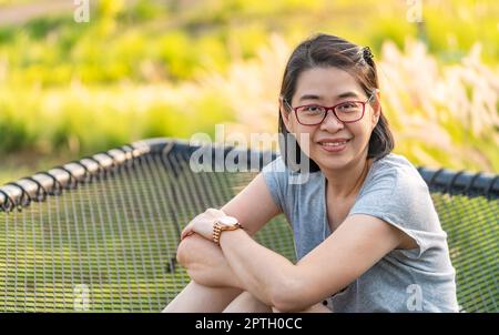 Porträt einer gesunden Frau mittleren Alters Entspannen Sie sich im Garten des Resorts, sitzen Sie auf einem Netzsitz, verschwommener Garten im Hintergrund, natürliches Sonnenlicht, Augen schauen auf Camer Stockfoto