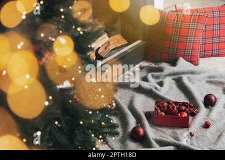 Karton mit Weihnachtsbällen, die auf grauem Teppich oder Karo liegen und zum Dekorieren und Aufhängen auf grüner Tanne mit Lichtern und Girlande im verwendet werden können Stockfoto