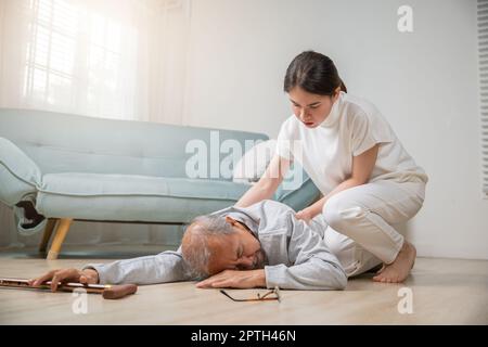 Der ältere asiatische Mann mit Gehstock fällt auf den Boden und die Enkeltochter wird im Wohnzimmer zu Hause unterstützt, während die junge Frau ihre großmutter halbieren wird Stockfoto