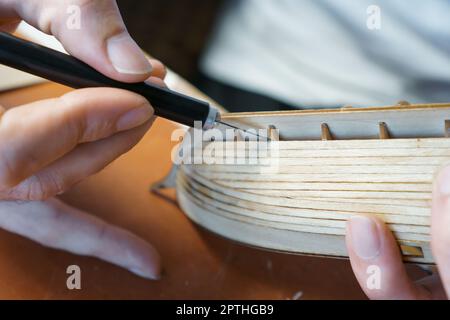 Hände des Mannes, der überschüssigen Klebstoff von Sperrholzdetails für Schiffsmodell mit Gebrauchsmesser entfernt. Prozess des Bauens Spielzeug Schiff, Hobby, Handwerk. Tabelle mit Stockfoto