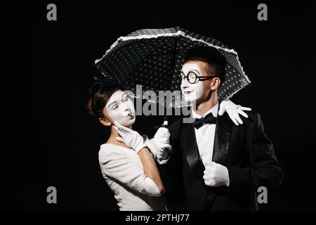 Portrait von Paar mime mit Sonnenschirm auf schwarzen Hintergrund. Mann im Smoking und Gläser und Frau in weißem Kleid Stockfoto