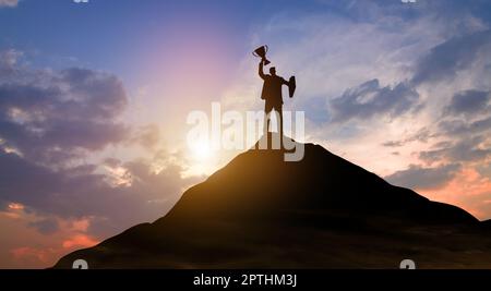 Silhouette Sieg Geschäftsmann mit Trophäe kletterte auf die Spitze des Berges über den Himmel und Sonnenlicht Hintergrund. Erfolg, Zielerreichung, Führung, Stockfoto