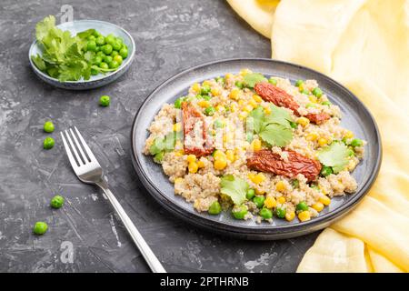 Quinoa-Haferbrei mit grüner Erbse, Mais und getrockneten Tomaten auf Keramikplatte auf grauem Betongrund und gelbem Textil. Seitenansicht, Nahaufnahme. Stockfoto