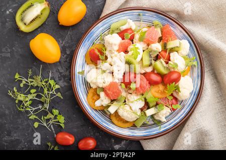 Vegetarischer Salat aus Blumenkohl, Kiwi, Tomaten, mikrogrünen Sprossen auf schwarzem Betongrund und Leinentextilien. Draufsicht, flach liegend, geschlossen Stockfoto