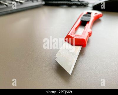 Schreibwaren auf dem Tisch im Büro. Schreibtischmesser mit rotem Kunststoffgriff. Papierschneider, Werkzeug für die Arbeit. Messer mit einer scharfen Metallklinge. Stockfoto