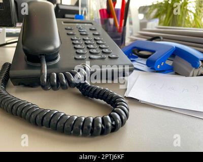 Schwarzes Festnetztelefon mit Röhre, Knöpfen und einem Kabel auf dem Arbeitstisch am Schreibtisch mit Büromaterial. Geschäftsarbeit. Stockfoto