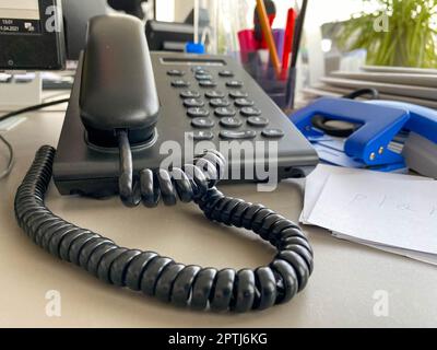 Schwarzes Festnetztelefon mit Röhre, Knöpfen und einem Kabel auf dem Arbeitstisch am Schreibtisch mit Büromaterial. Geschäftsarbeit. Stockfoto