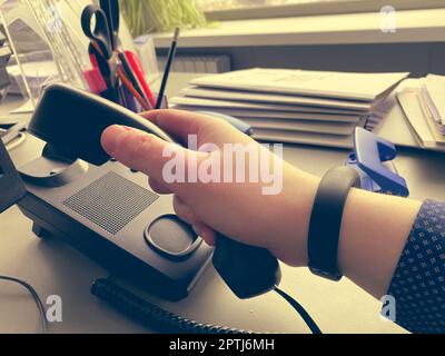 Die Hand eines Mannes in einem Hemd hält einen Festnetz-Telefonhörer auf einem Schreibtisch mit Büromaterial in einem Geschäftsbüro. Stockfoto