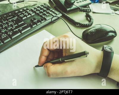 In der Hand des Büromitarbeiters befindet sich ein schwarzer Metallstift mit einer Stange. Handschriftliche Notizen auf einem weißen Stück Papier machen. Ein Mann, der neben einem Computer schreibt Stockfoto