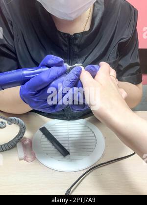 Die Frau, das Mädchen, der Meister in der Maniküre, ein Nagelservice, macht eine Maniküre in dunkelblauen Handschuhen an Händen das Gerät eine Nagelfeile. Stockfoto