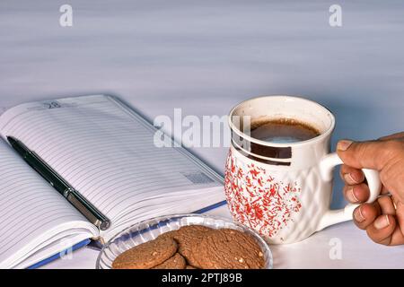 Eine Hand nimmt eine Tasse schwarzen Tee vom Arbeitstisch, um sie zusammen mit einem Teller Kekse und einem Tagebuch mit Stift zu genießen - ideal für Werbung Stockfoto