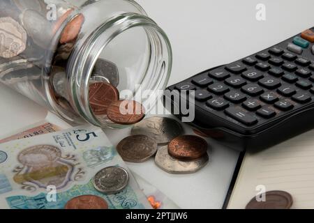 Glasflasche umgefallen, verschüttet Münzen auf weißen Tisch mit Banknoten, Rechner, Notizbuch und Stift Stockfoto