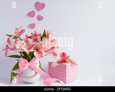 Strauß rosa Blumen in einer kleinen weißen Vase und ein Geschenk daneben mit Papier mit kleinen rosa Herzen und einer Blume auf den Blumen, die Sie sehen können Stockfoto