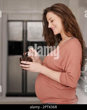 Sich ihrem Schwangerschaftsverlangen hingeben. Eine schwangere Frau, die einen Cupcake in der Küche isst Stockfoto