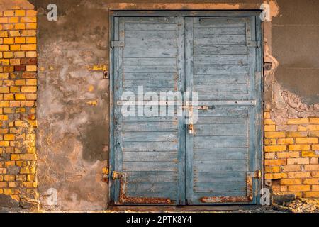 Alte geschlossene Tür mit Vorhängeschloss, vor dem Hintergrund einer alten, abblätternden Wand. Ziegel und Risse an der Wand. Vorderansicht. Stockfoto