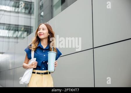 Asiatische schöne Geschäftsfrau zuversichtlich lächelnd mit Stofftasche hält Stahl Thermoskannen Becher Wasser Glas sie zu Fuß im Freien auf der Straße in der Nähe Modus Stockfoto