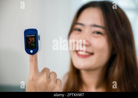 Asiatische junge Frau, die medizinische Oxymeter-Ausrüstung verwendet, um Puls-Sauerstoffsättigung auf dem Sofa zu Hause im Wohnzimmer zu messen, Self Healthcare mit Corona viru Stockfoto