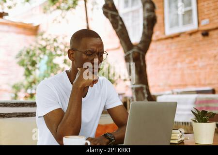Ein afroamerikanischer Mann mit Brille, der am Tisch im Café auf der Sommerveranda in Gedanken saß, über Ideen nachdachte und Probleme in der Nahaufnahme löste. Ich arbeite online an Stockfoto