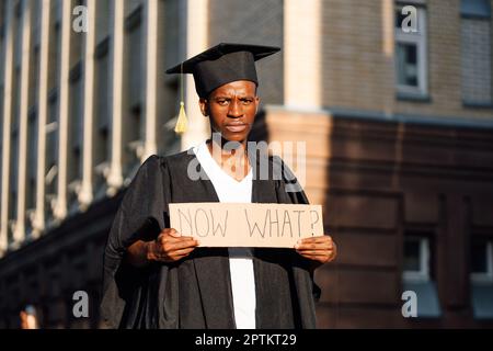 Porträt eines verärgerten afroamerikanischen Mannes, der mit einem Pappposter auf der Straße neben dem Gebäude auf der Suche nach einem Job stand. Universitäts- oder Hochschulabschluss Stockfoto