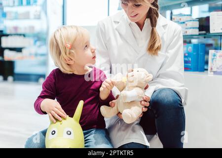 Kleines Mädchen in der Apotheke im Gespräch mit der Apotheke sitzt auf einem Spielzeug Stockfoto