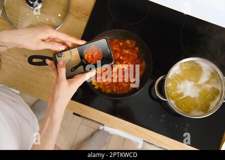 Blick von oben auf eine nicht erkennbare Frau mit Smartphone, die beim Kochen in der Küche im hom eine Bratpfanne mit Tomatensauce für einen kulinarischen Blog fotografiert Stockfoto