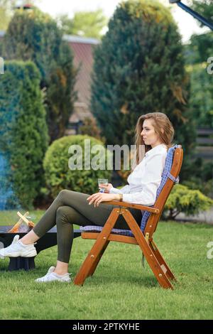 Junge Frau sitzt im Liegestuhl entspannt auf dem Hinterhof neben einem Grill, im Profil ansehen Stockfoto