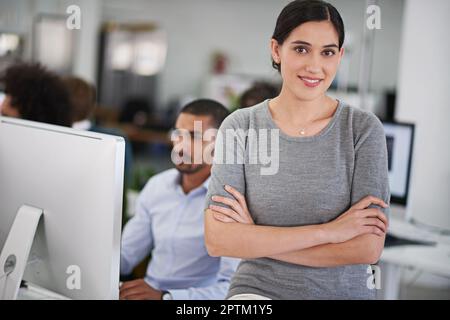 Shes bekam Geschäft Akkumene. Eine attraktive junge Geschäftsfrau sitzt auf ihrem Schreibtisch mit ein paar Kollegen im Hintergrund Stockfoto