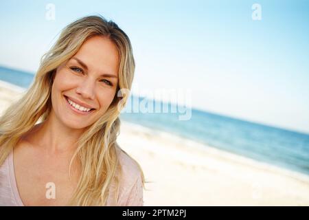 Das ist das Paradies. Kopf- und Schulterporträt einer attraktiven jungen Frau, die an einem sonnigen Strand steht Stockfoto