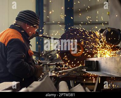 Ein weißer Mann arbeitet an der Maschine, schneidet Metall, die Arbeiten werden in der Werkstatt durchgeführt, er trägt einen Overall, Funken fliegen aus der Maschine in alle Richtungen. Stockfoto