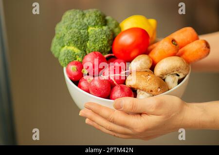 In den Händen einer weißen Frau, eine milchige Keramikschüssel voller Gemüse: Brokkoli, rote Rettiche, eine Zitrone, Tomaten, Schweinefilze und sogar Karotten Stockfoto