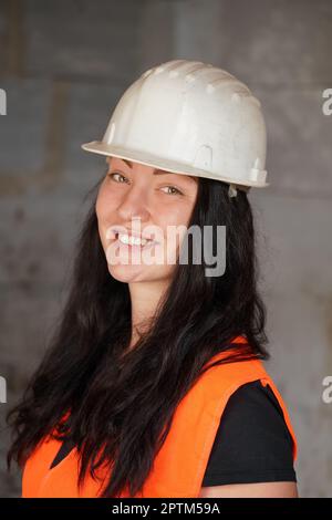 Junge Frau in weißem Schutzhelm und orangefarbener Warnjacke, langes dunkles Haar, schaut in die Kamera und lächelt. Nahaufnahme von Detail zu Gesicht, verschwommene Kontras Stockfoto