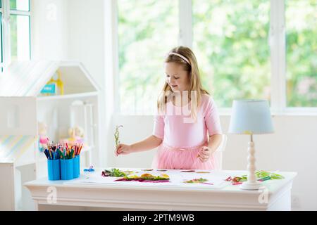 Kind schafft Bild mit bunten Blättern. Kunst und Kunsthandwerk für Kinder. Kleines Mädchen macht Collage Bild mit Regenbogenpflanzen Blatt. Hausaufgaben für die Biologie Stockfoto