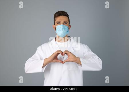 Arzt in Schutzmaske macht Herz mit Händen auf grauem Hintergrund Stockfoto