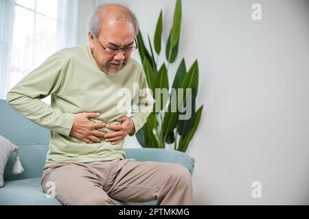 Älterer asiatischer alter Mann, der auf dem Sofa sitzt, Magenschmerzen hat, im Alter Magenschmerzen hat und seinen Magen im Wohnzimmer hält, die Gesundheit der Menschen ist sehr gesund Stockfoto
