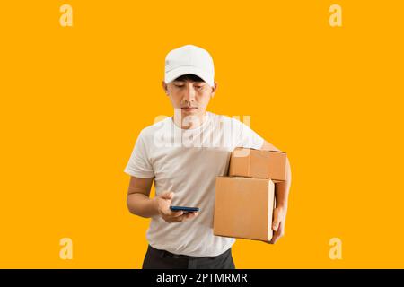 Junge asiatische Lieferung Mann trägt Kappe mit weißen leeren T-Shirt hält Smartphone und Paket-Briefkasten isoliert auf gelbem Hintergrund. express-Lieferung s Stockfoto