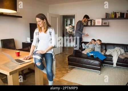 Schwangere Frau mit Laptop auf dem Esstisch, während der Vater den Söhnen im Hintergrund in München Saft anbietet Stockfoto