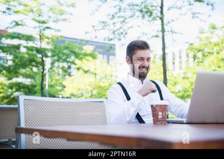 Geschäftsmann, der an einem Laptop arbeitet, in einem Café im Freien und lächelnd, München, Bayern, Deutschland Stockfoto