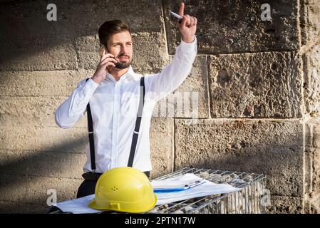 Männlicher Architekt, der mit einem Bauplan auf einer Baustelle in München, Bayern, spricht Stockfoto