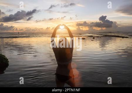 Silhouette einer Frau, die bei Sonnenuntergang über dem Ozean, Uluwatu, Bali, Indonesien ihr Herz Formen lässt Stockfoto