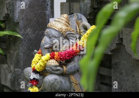 Traditionelle Ganesha-Statue mit Blumenkranz im balinesischen Garten. Lord Ganesha soll Glück bringen, und so wird er vor allem verehrt Stockfoto