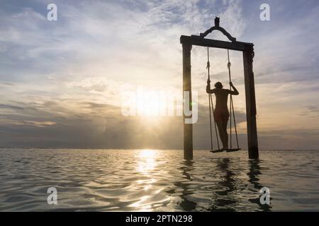 Rückansicht eines jungen Mannes, der auf einer Seilschaukel am Strand gegen Sonnenuntergang steht, Gili Trawangan, Lombok, Indonesien Stockfoto