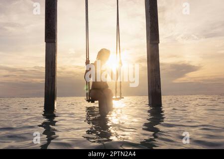 Eine Frau, die auf einem Seil am Strand gegen Sonnenuntergang sitzt, Gili Trawangan, Lombok, Indonesien Stockfoto